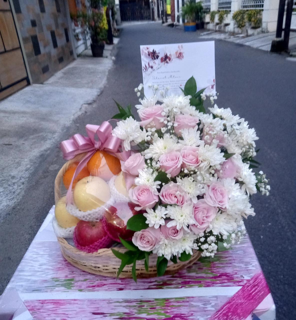 Parcel bunga mawar pink chrysanthemum putih dan buah dalam keranjang anyaman dengan pita pink untuk ucapan selamat melahirkan
